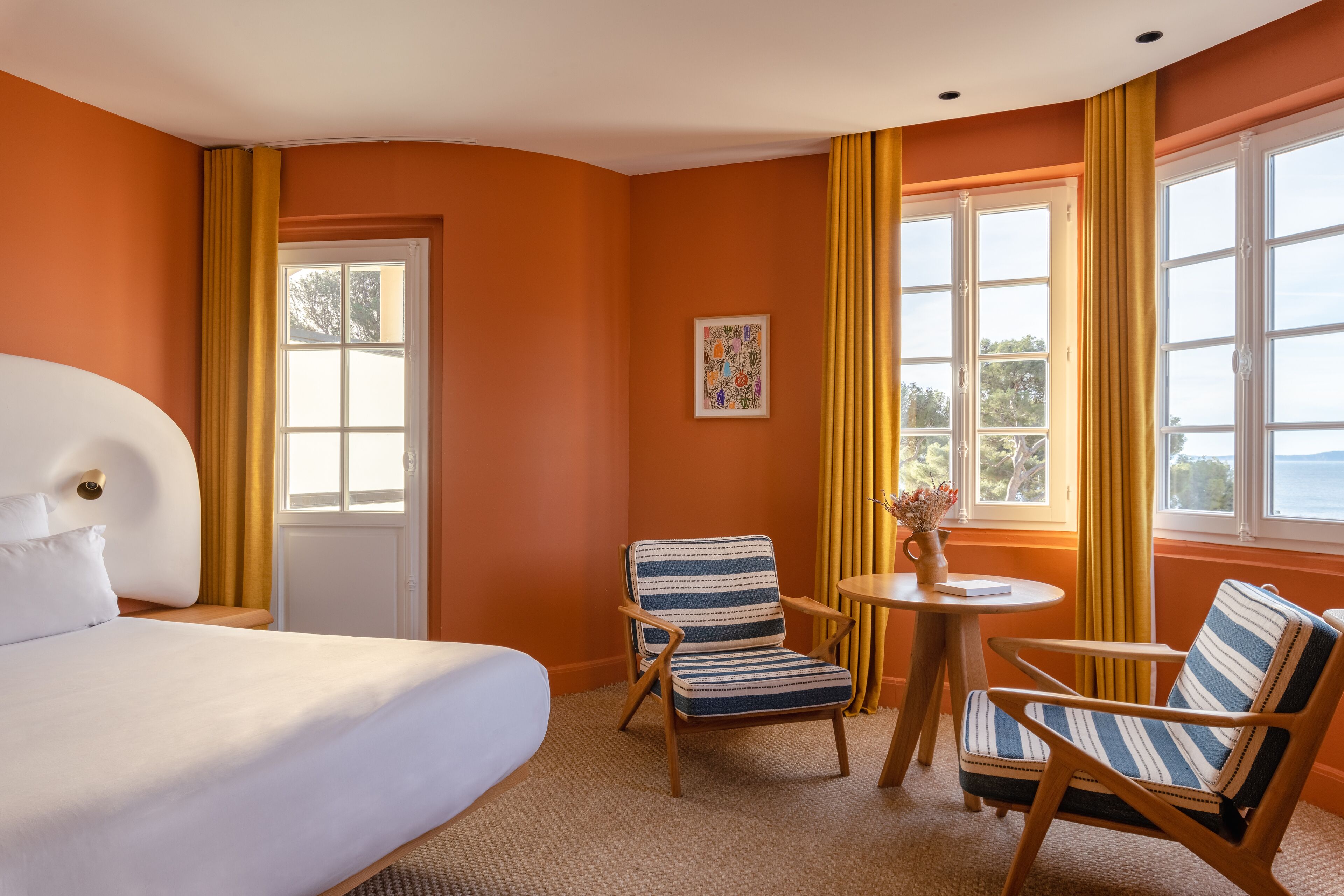 Een hotelkamer met een groot bed, twee gestreepte fauteuils, een klein rond tafeltje en een raam met gordijnen.