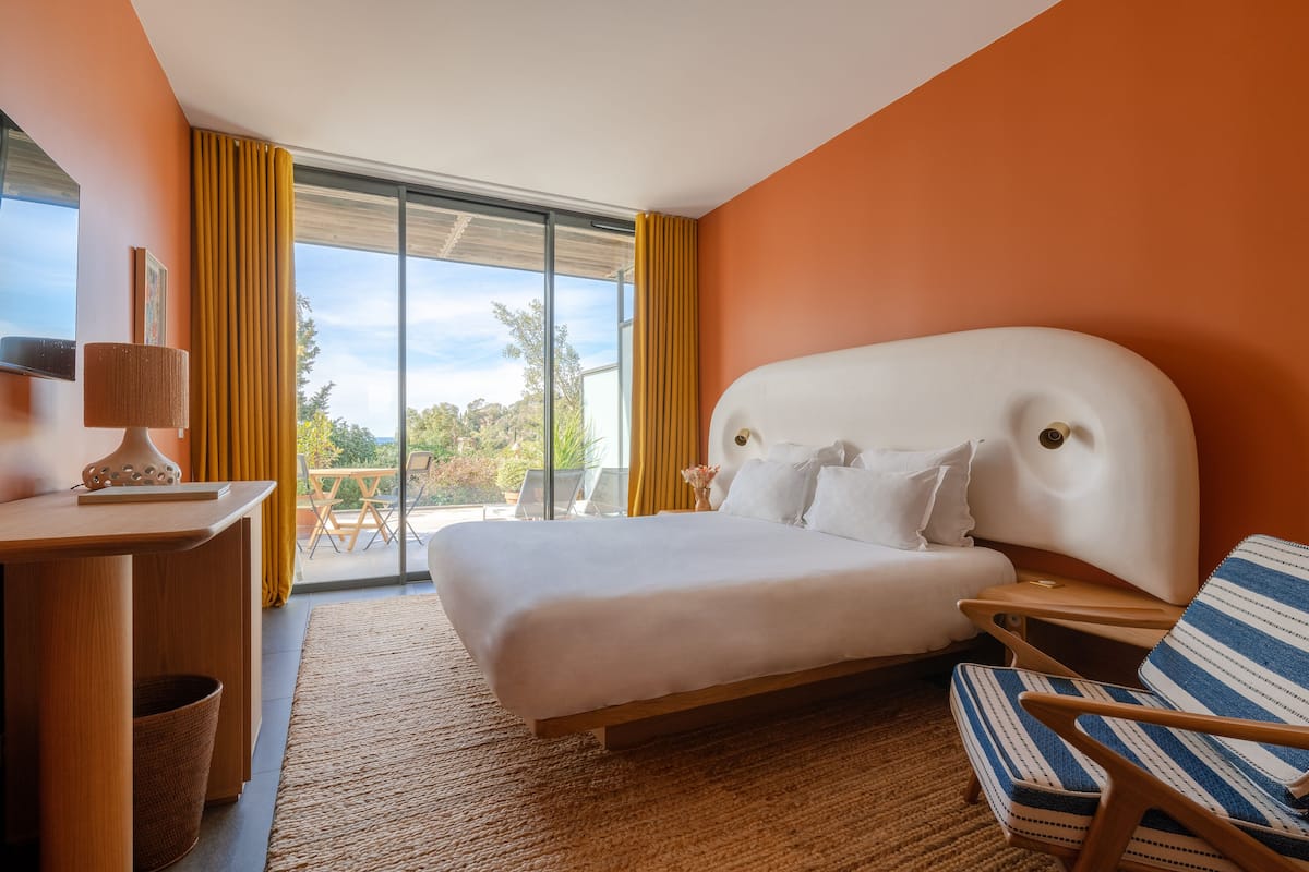 A modern hotel room with a large bed, a wooden desk, a striped chair, and a view of greenery through large windows.