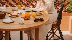 Une femme est assise à une table en plein air, devant un petit-déjeuner, et elle jouit d’une vue panoramique sur un paysage verdoyant, avec des arbres et la mer à l’horizon.