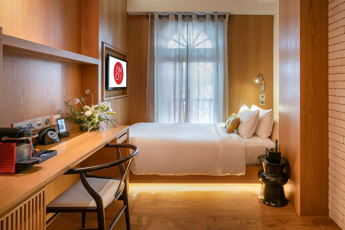 A hotel room with a wooden desk, a bed with white linens, a small table with a vase of flowers, and a wall-mounted TV.