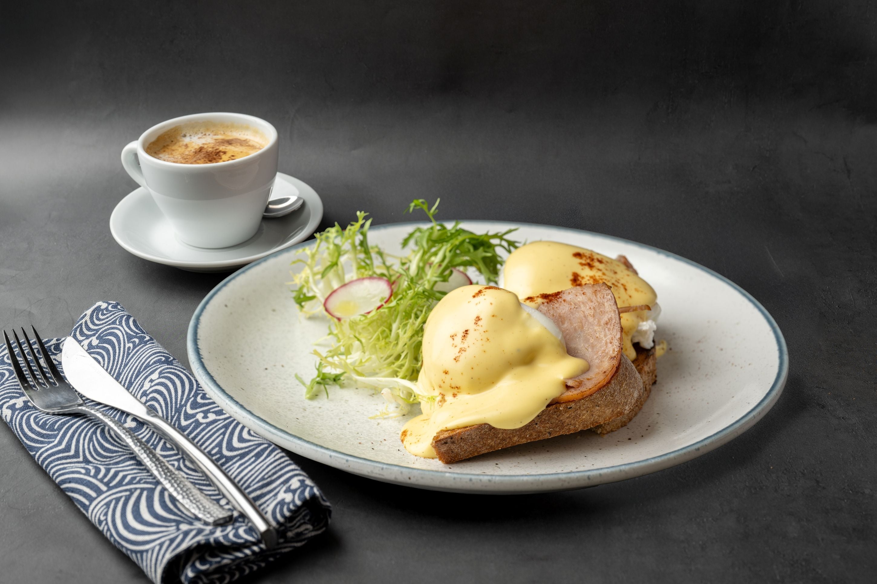 Un plato con huevos Benedictinos, una taza de café y una servilleta con un tenedor sobre una superficie oscura.