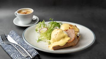 Un plato con huevos Benedictinos, una taza de café y una servilleta con un tenedor sobre una superficie oscura.