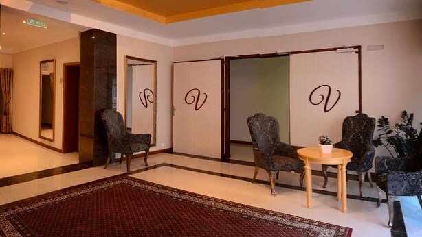 Een hotel lobby met een rood tapijt, donkere fauteuils, een ronde houten tafel en een grote spiegel met decoratieve lijst.