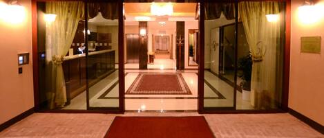 A hotel lobby with a red carpet, glass doors with curtains, a reception desk, and a chandelier.