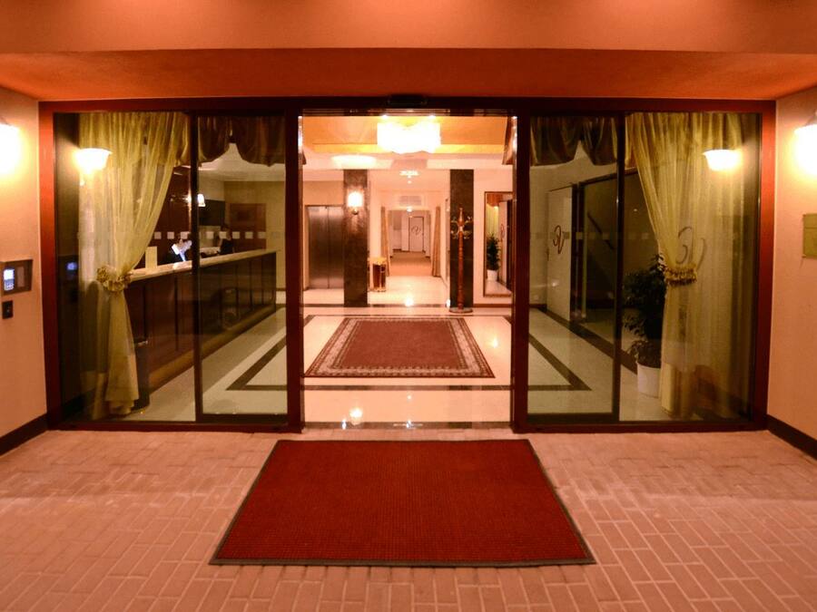 Un vestíbulo de hotel con alfombra roja, puertas de cristal con cortinas, un mostrador de recepción y una araña.