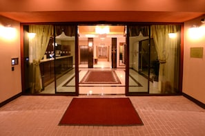 Un vestíbulo de hotel con alfombra roja, puertas de cristal con cortinas, un mostrador de recepción y una araña.