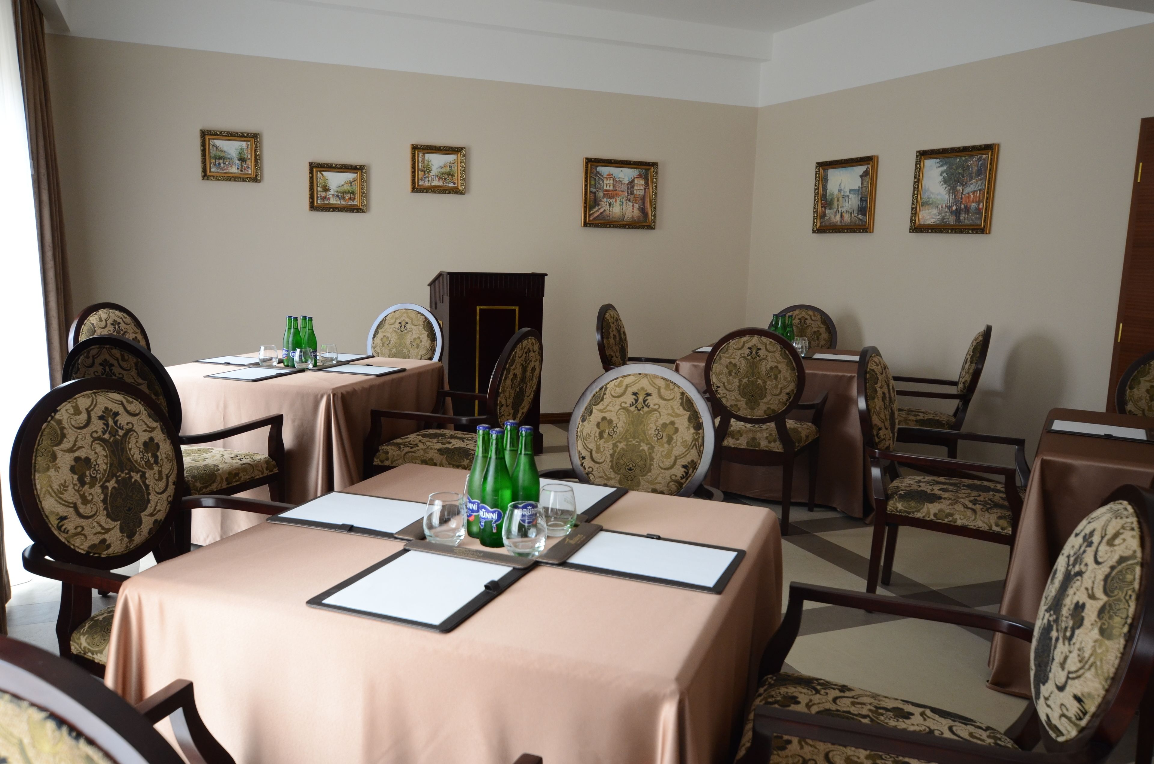 Ein Essbereich mit Tischen für vier Personen, jeder Tisch mit einer beigen Tischdecke, Stühle mit gemusterten Bezügen und grüne Flaschen auf den Tischen.