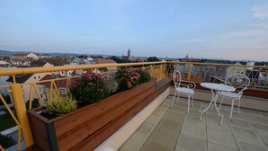 Un balcón con banca de madera, sillas de metal blanco y barandilla amarilla con vistas a la ciudad.