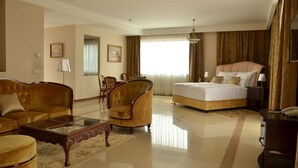 Habitación de hotel con una cama grande, dos sillones, un sofá, una mesa de centro de vidrio y un escritorio con teléfono.