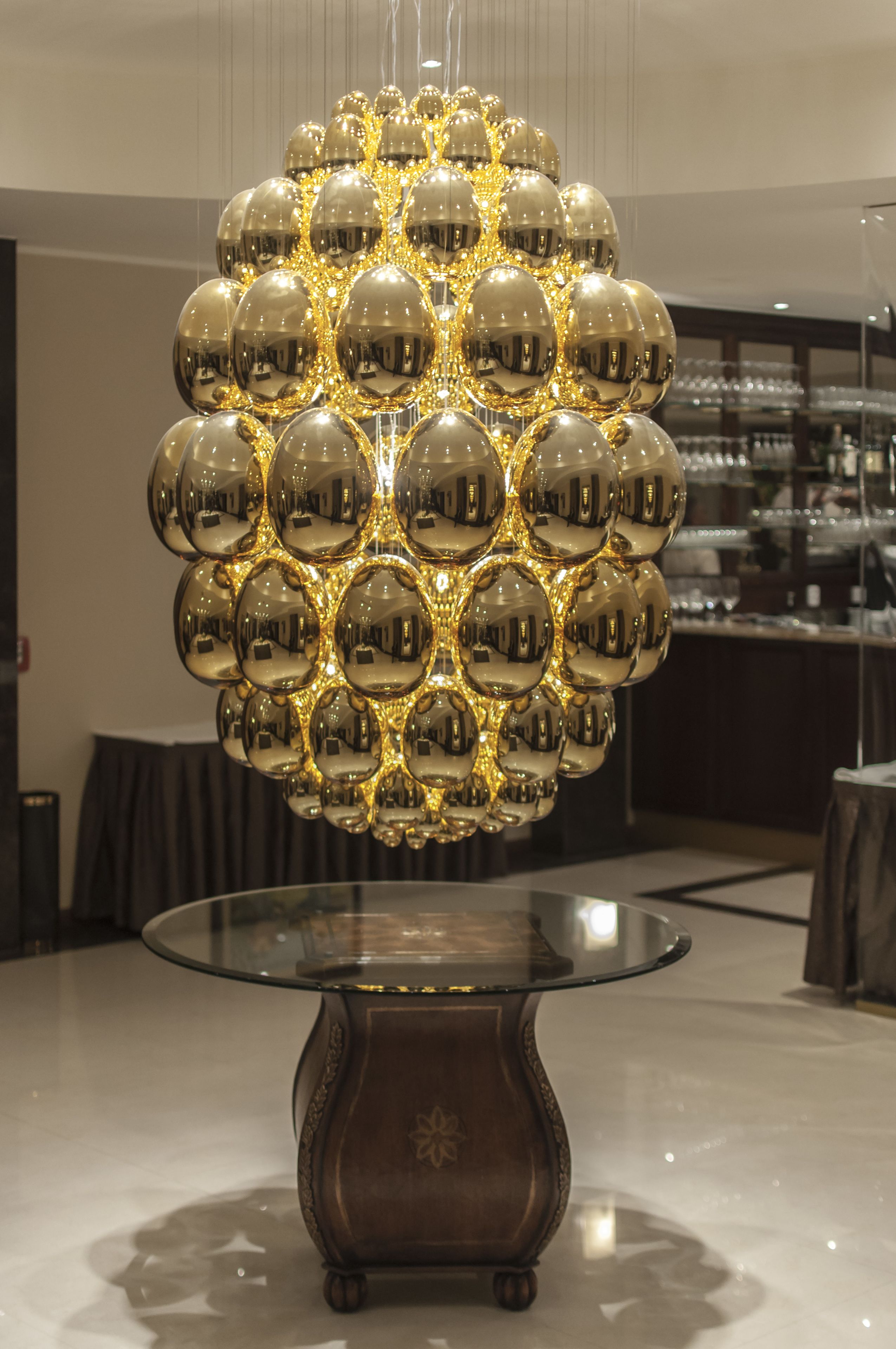 Una lámpara esférica con múltiples superficies reflectantes, una mesa de cristal con borde curvo y una base de madera oscura.