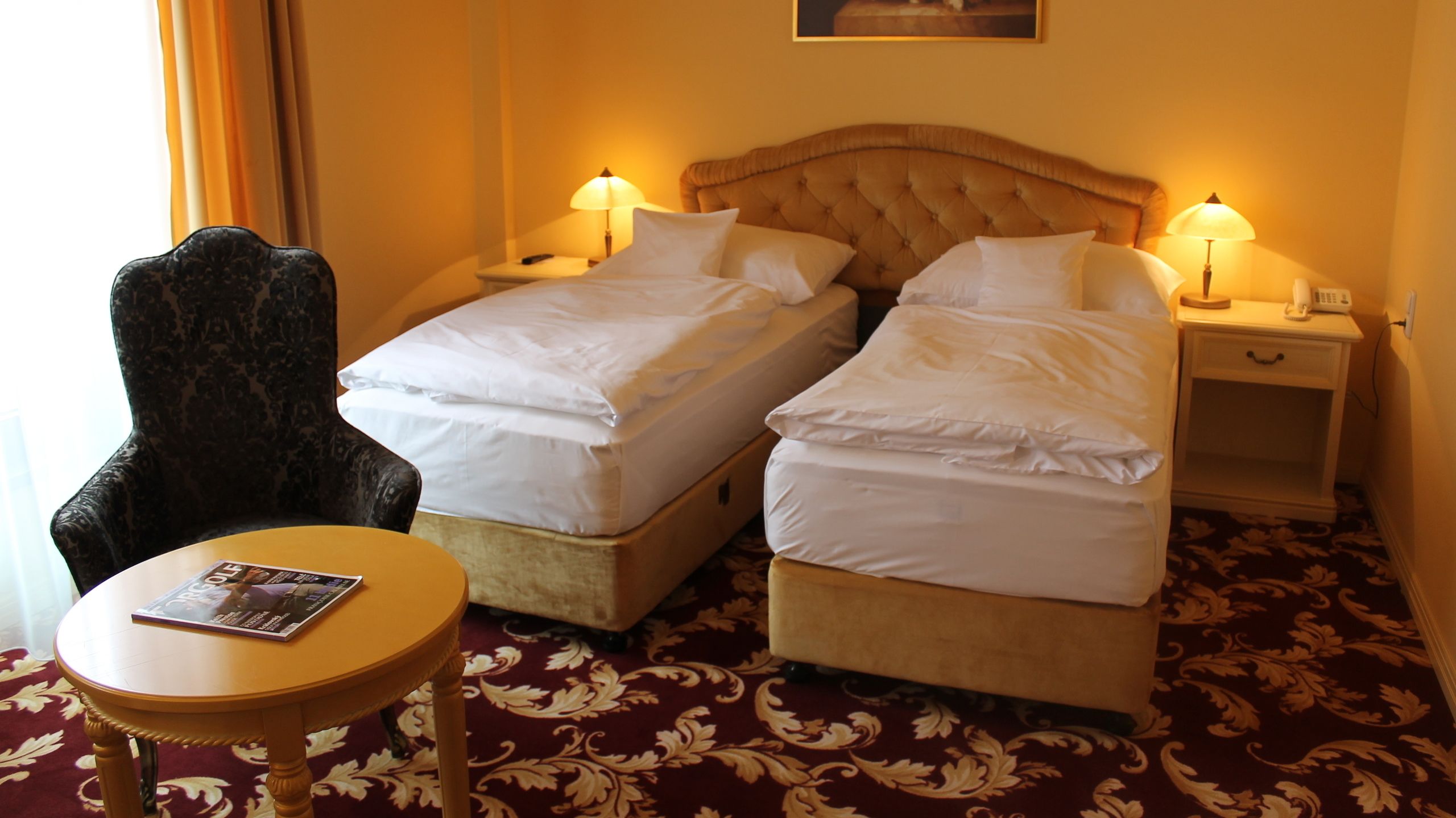 A hotel room with two beds, a chair, a table, and a nightstand with a lamp.