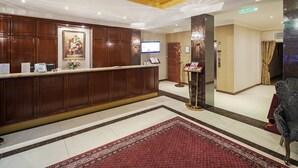Un vestíbulo de hotel con mostrador de recepción, paneles de madera, una araña, un televisor y una alfombra roja estampada.