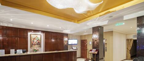 Un hall d’hôtel avec un comptoir d’accueil, des boiseries, un lustre, un téléviseur et un tapis rouge à motifs.