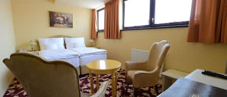 A hotel room with a bed, two chairs, a small table, a lamp, a painting on the wall, and a window with curtains.