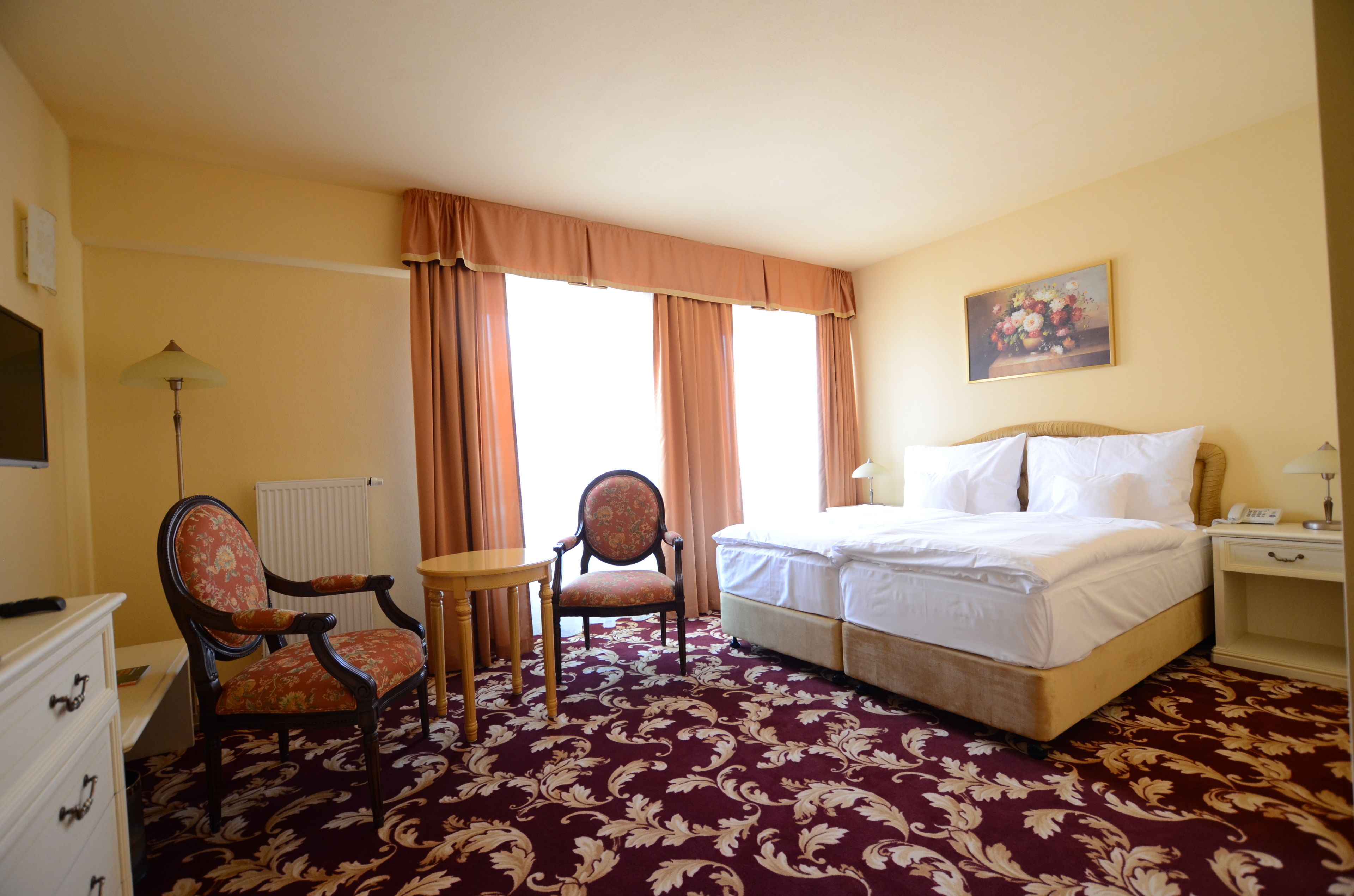 Una habitación de hotel con cama, dos sillas, una mesa, un radiador y un cuadro en la pared.