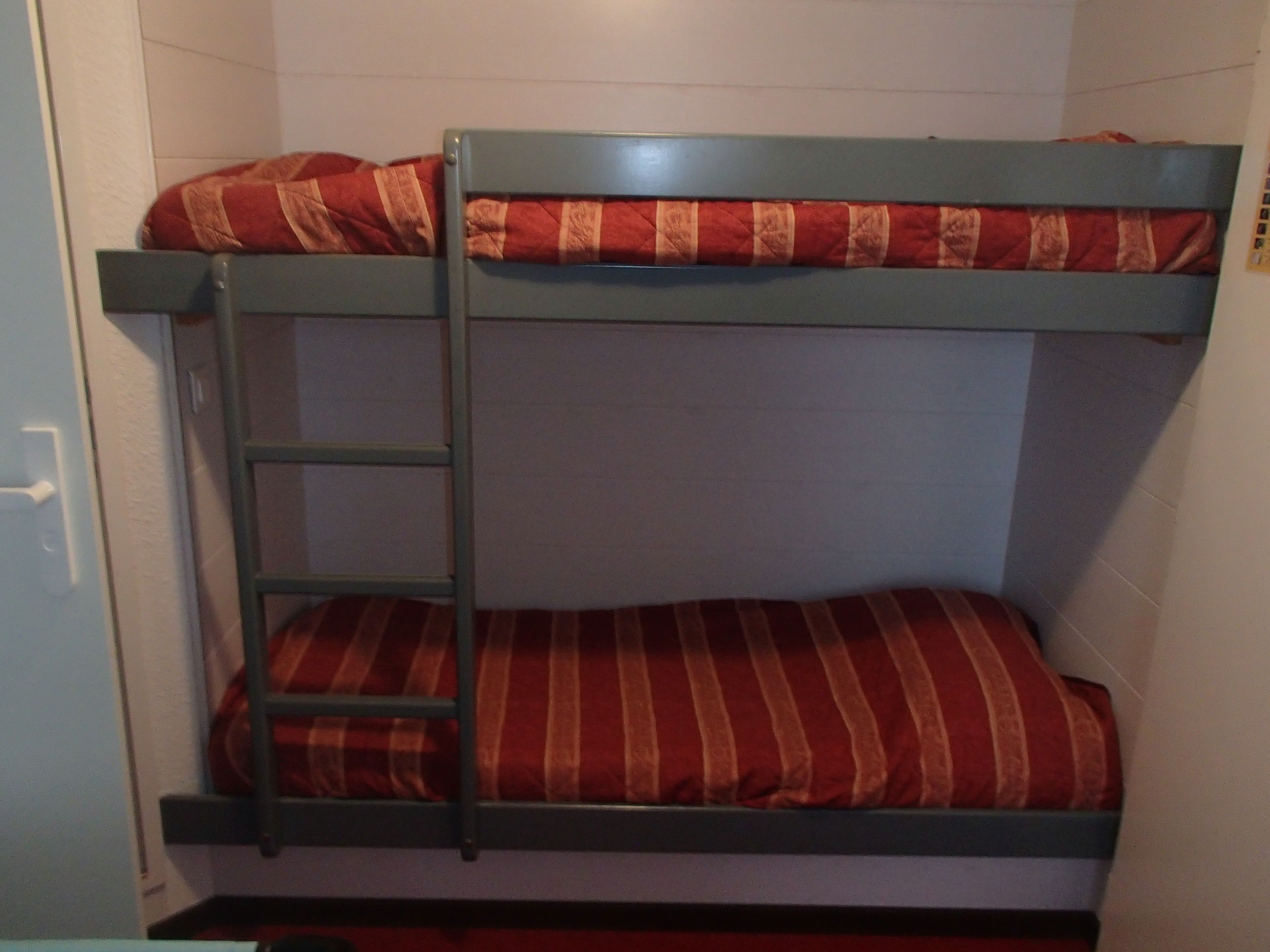 Una cama litera con escalera, ropa de cama a rayas rojas y blancas, y una pared blanca.