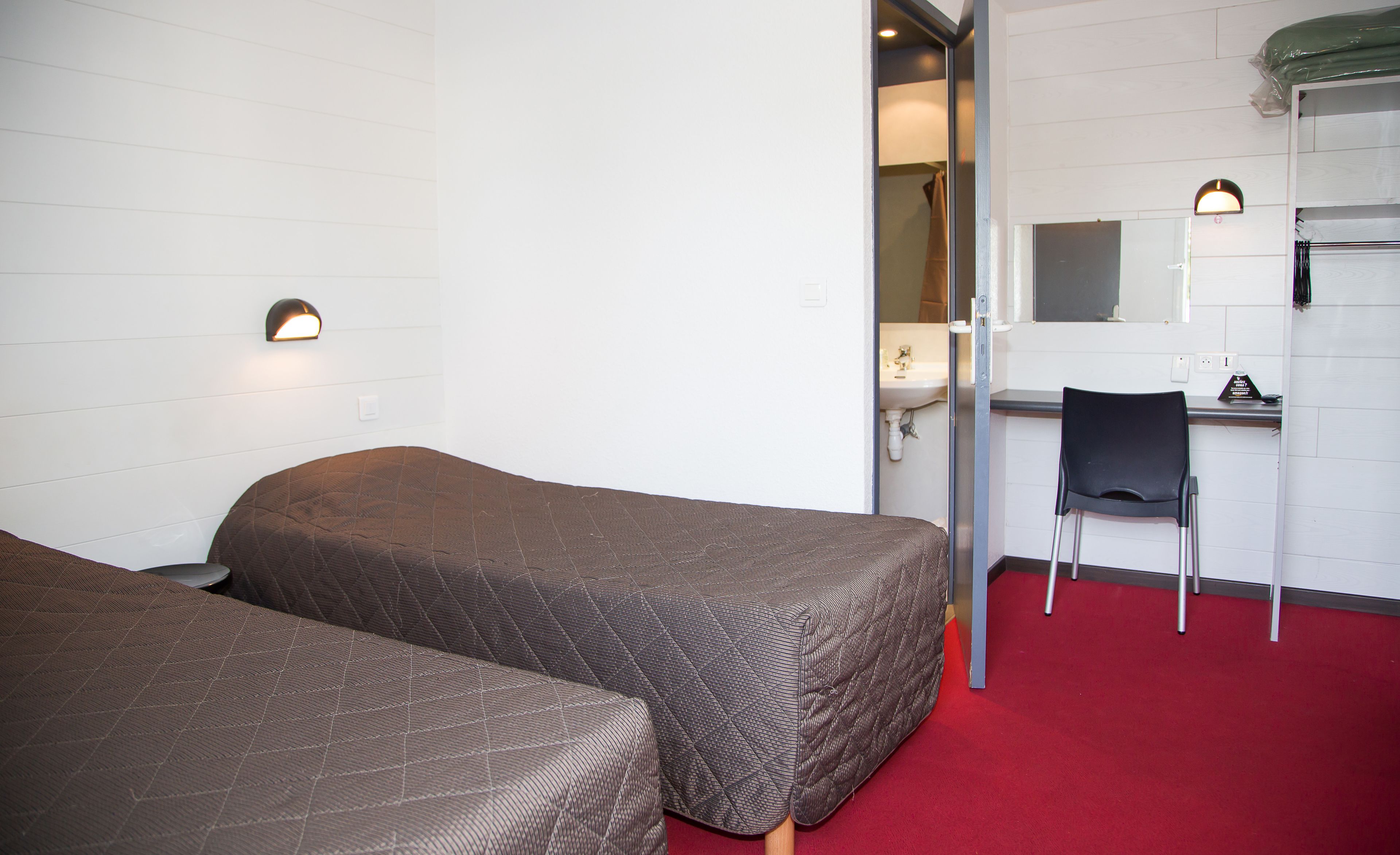 A small hotel room with two beds, a desk, a chair, a mirror, and a red carpet.