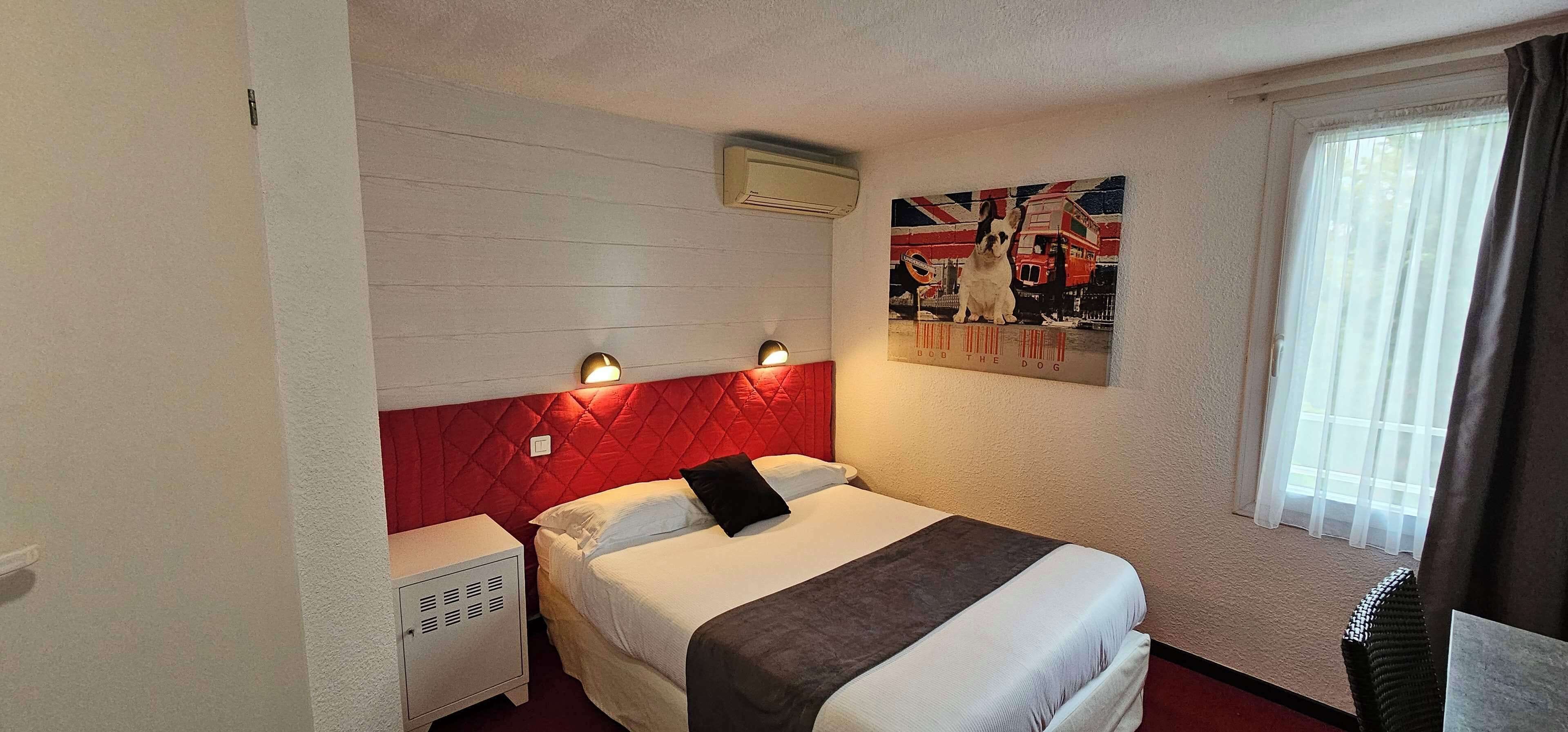Hotelkamer met een rode hoofdbord, een bed met witte lakens, een klein nachtkastje, een wandairconditioner en een poster met een hond op een bus.