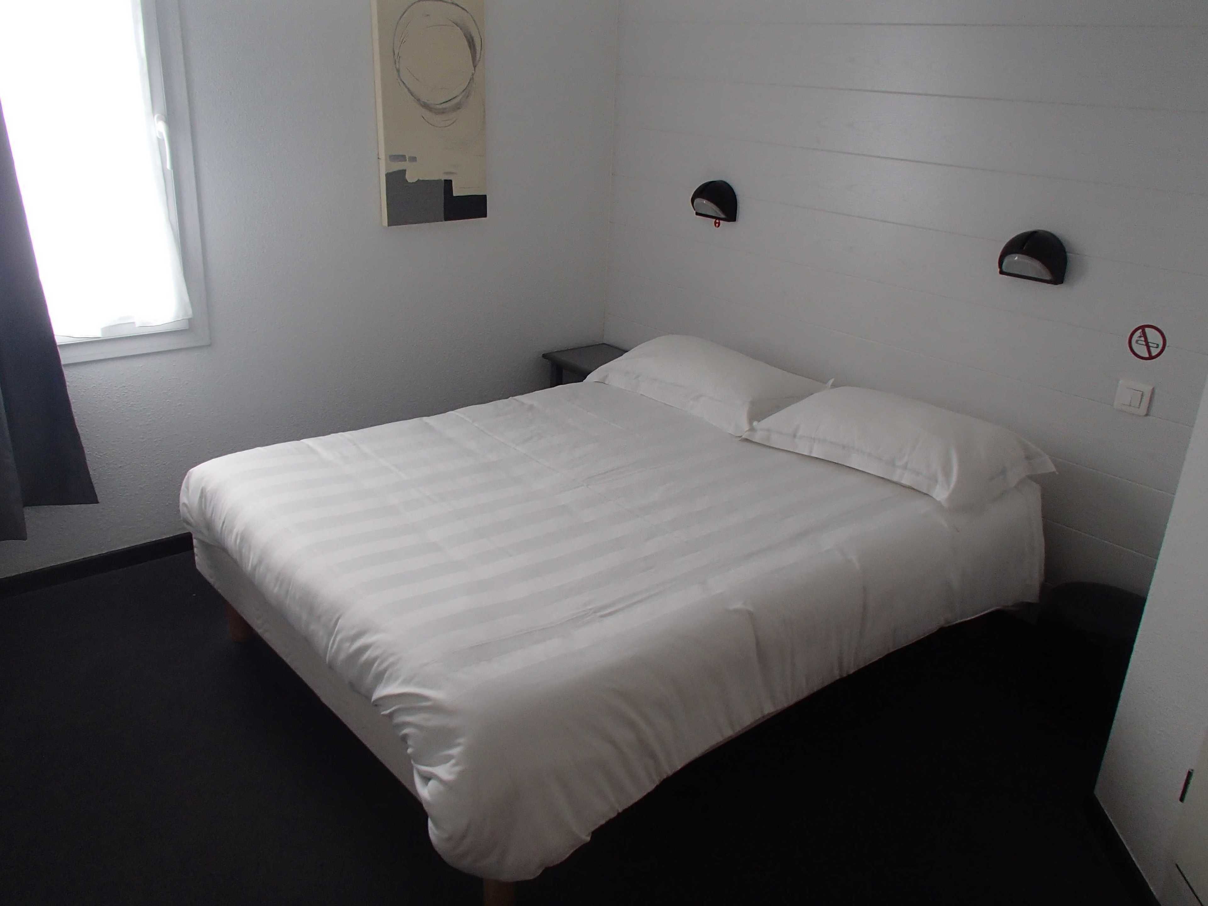 A neatly made bed with white bedding in a room with a window, a wall-mounted light fixture, and a no-smoking sign.