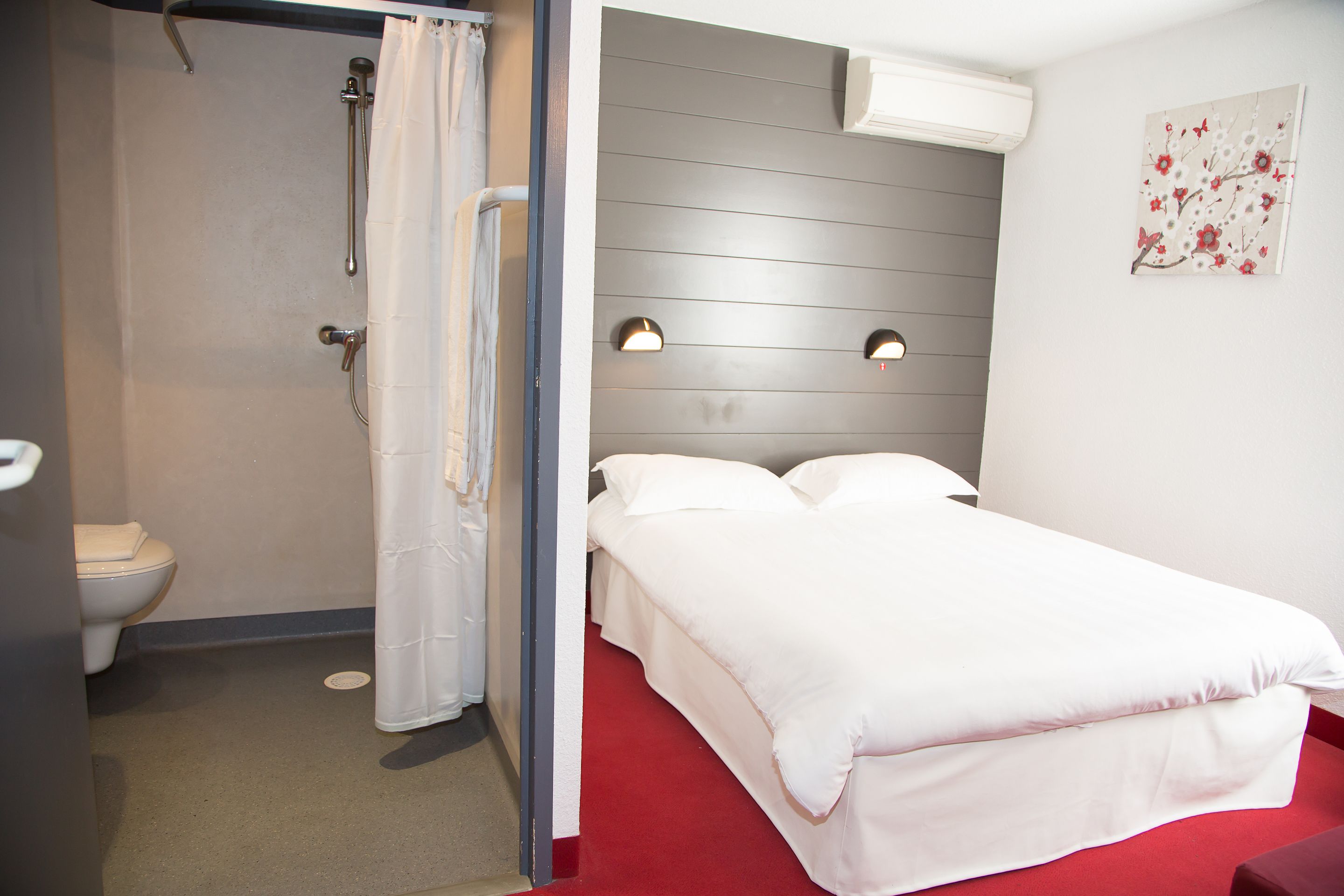 Habitación de hotel con ducha, inodoro, cama y dos luces fijadas a la pared.