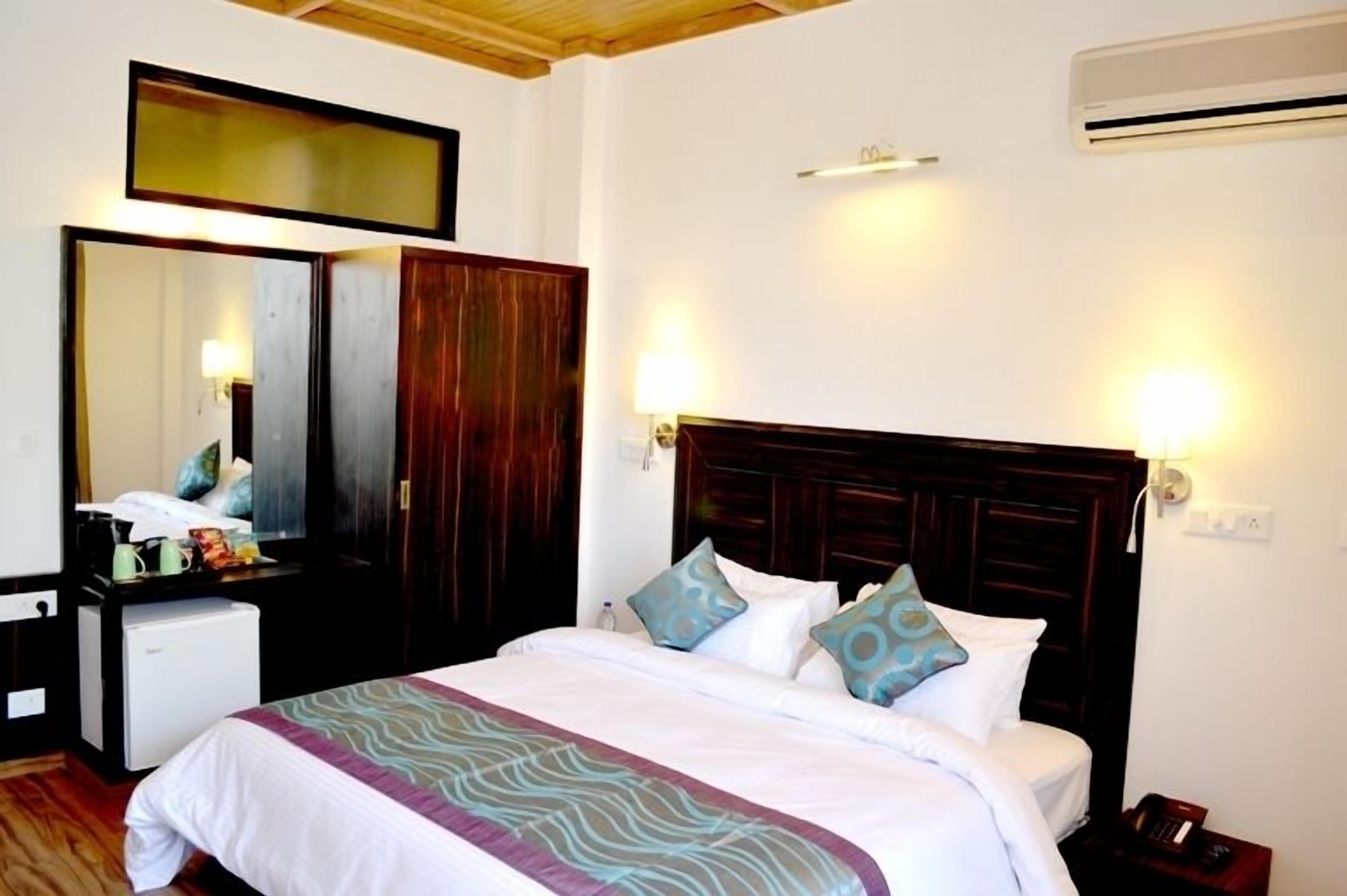 A hotel room with a bed, bedside lamps, a mirror, a wardrobe, a desk with a lamp, and an air conditioning unit.