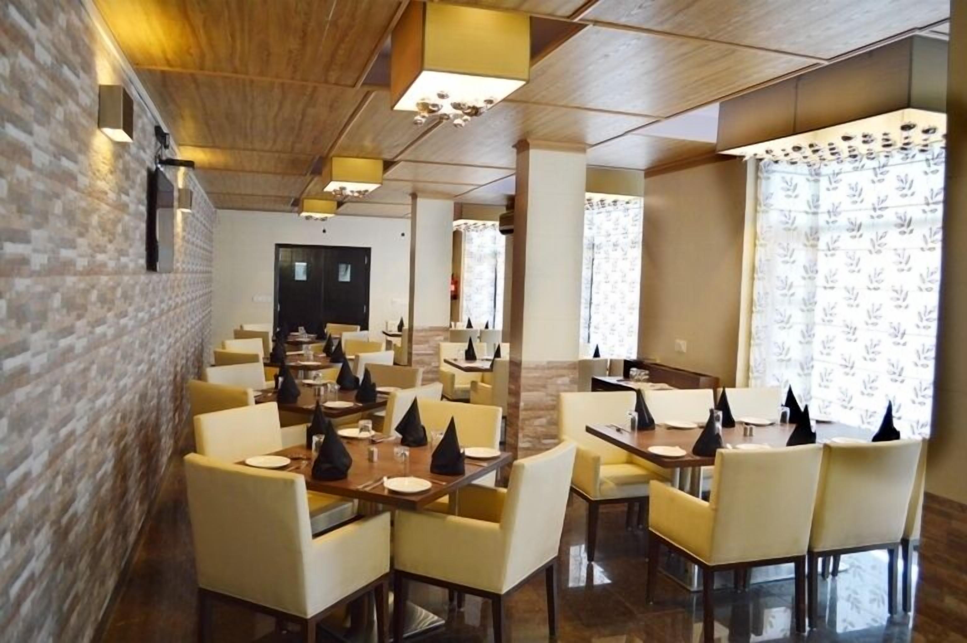A restaurant with multiple tables and chairs, wooden ceiling, brick wall, and patterned curtains.