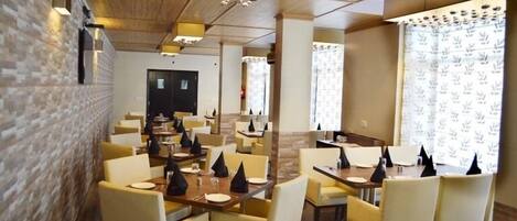 A restaurant with multiple tables and chairs, wooden ceiling, brick wall, and patterned curtains.
