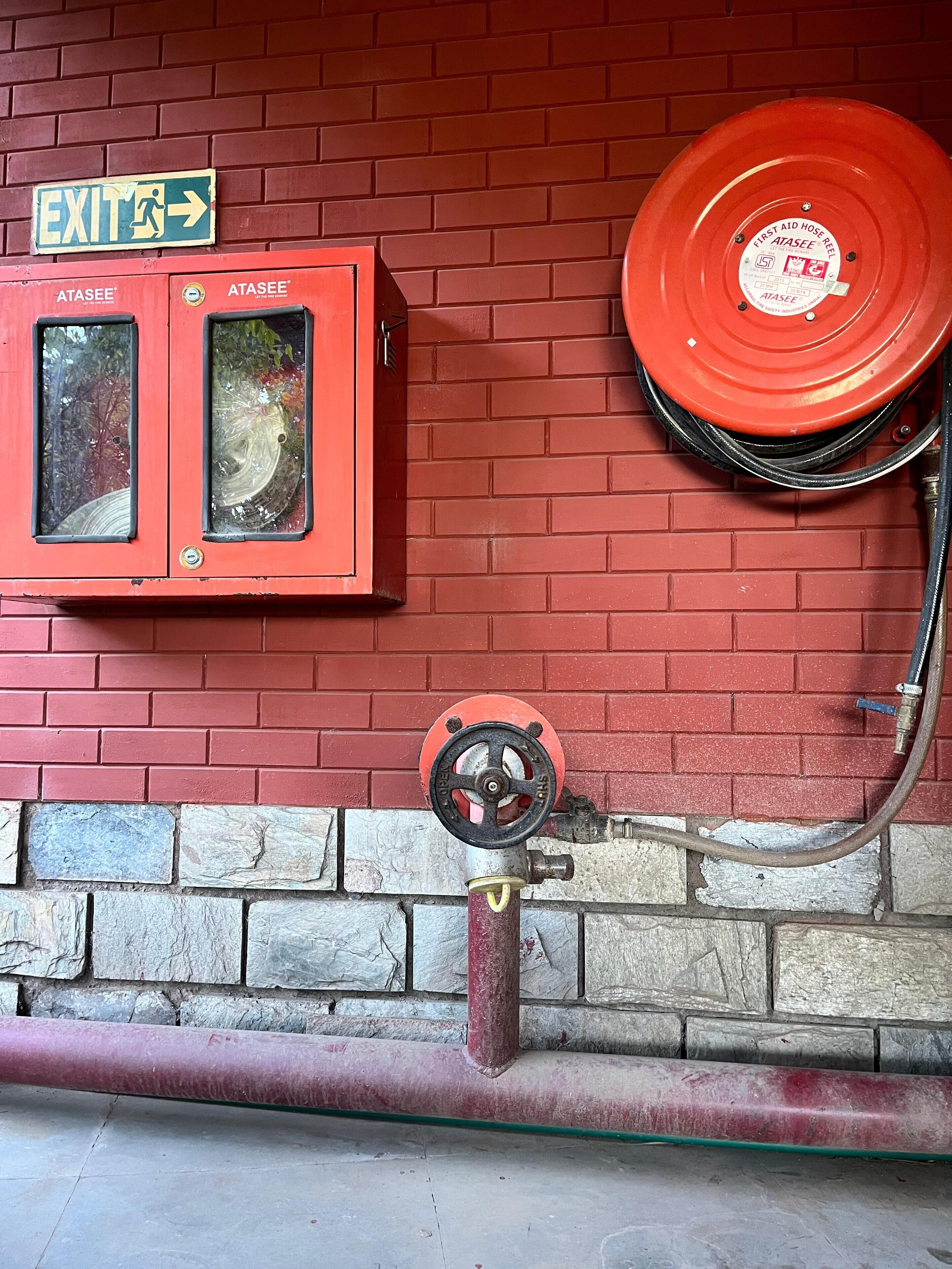 A red fire hose reel mounted on a brick wall, an exit sign, and two ATASEE fire extinguishers.