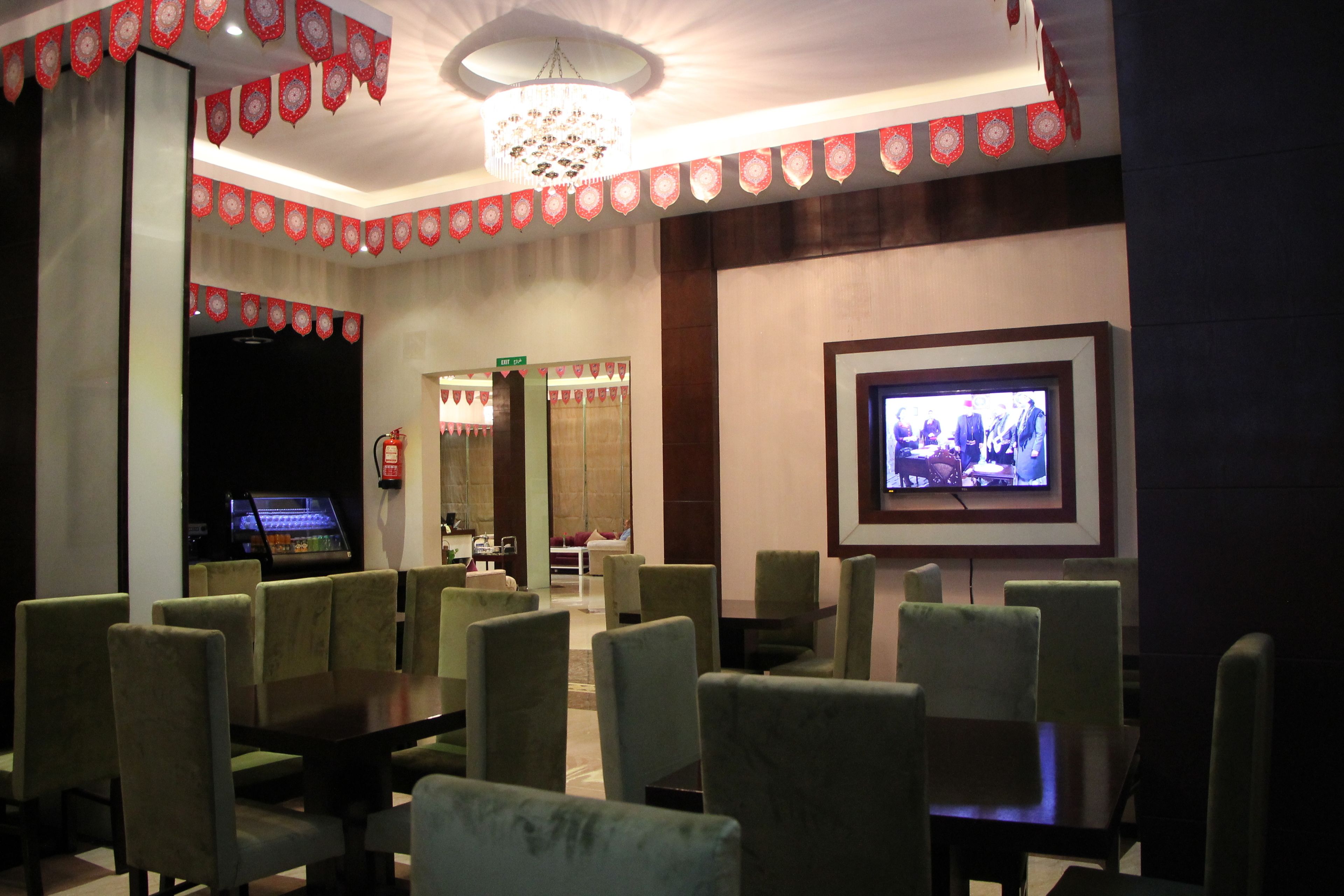 Un hall de hotel con un televisor montado en la pared, una araña y una zona de estar con sillones tapizados en verde y mesas.
