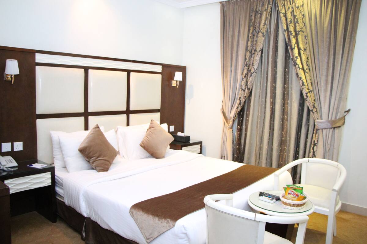 Une chambre d’hôtel avec un grand lit, une table de chevet avec une lampe, une chaise et une petite table sur laquelle se trouvent un panier et une télécommande.
