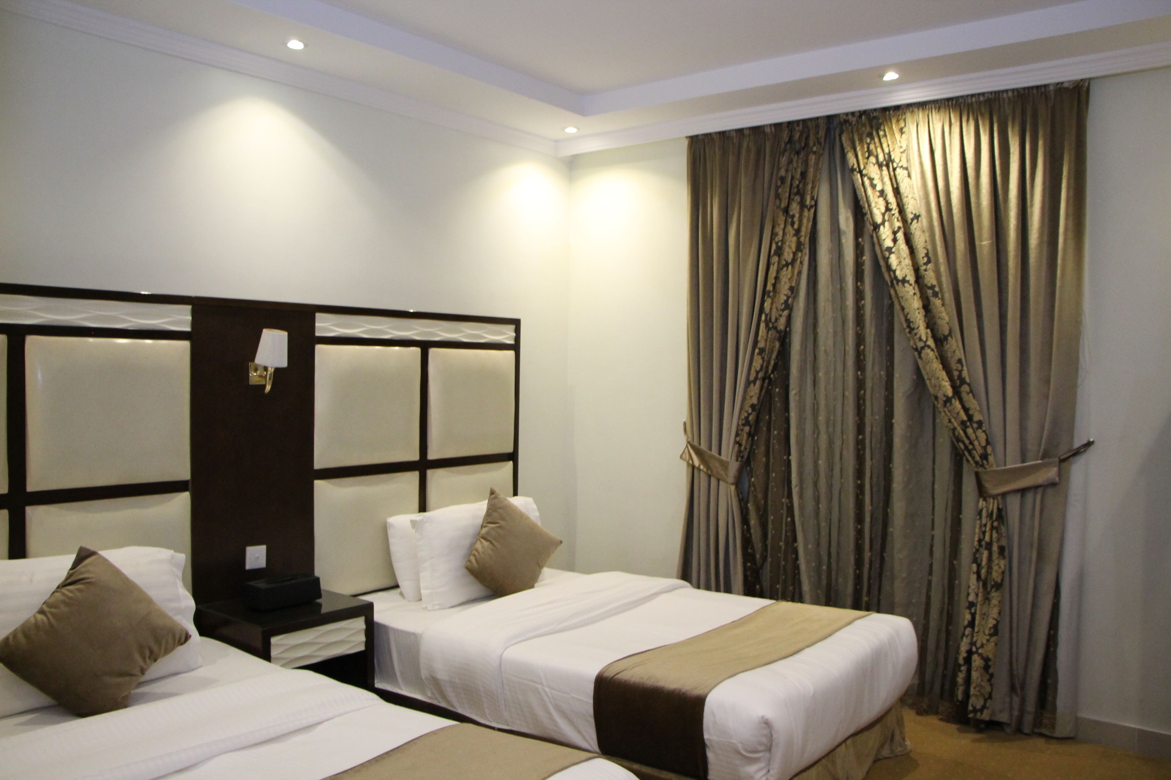 A hotel room with two beds, a nightstand, a lamp, and a large window with curtains.