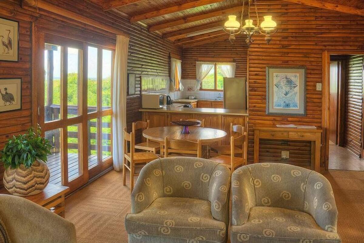 A rustic cabin interior with a dining area, kitchen, and large windows offering a view of greenery.