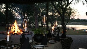 An outdoor dining area with tables set for a meal, surrounded by trees and a body of water in the background. - Bonamanzi Game Reserve (Hluhluwe)