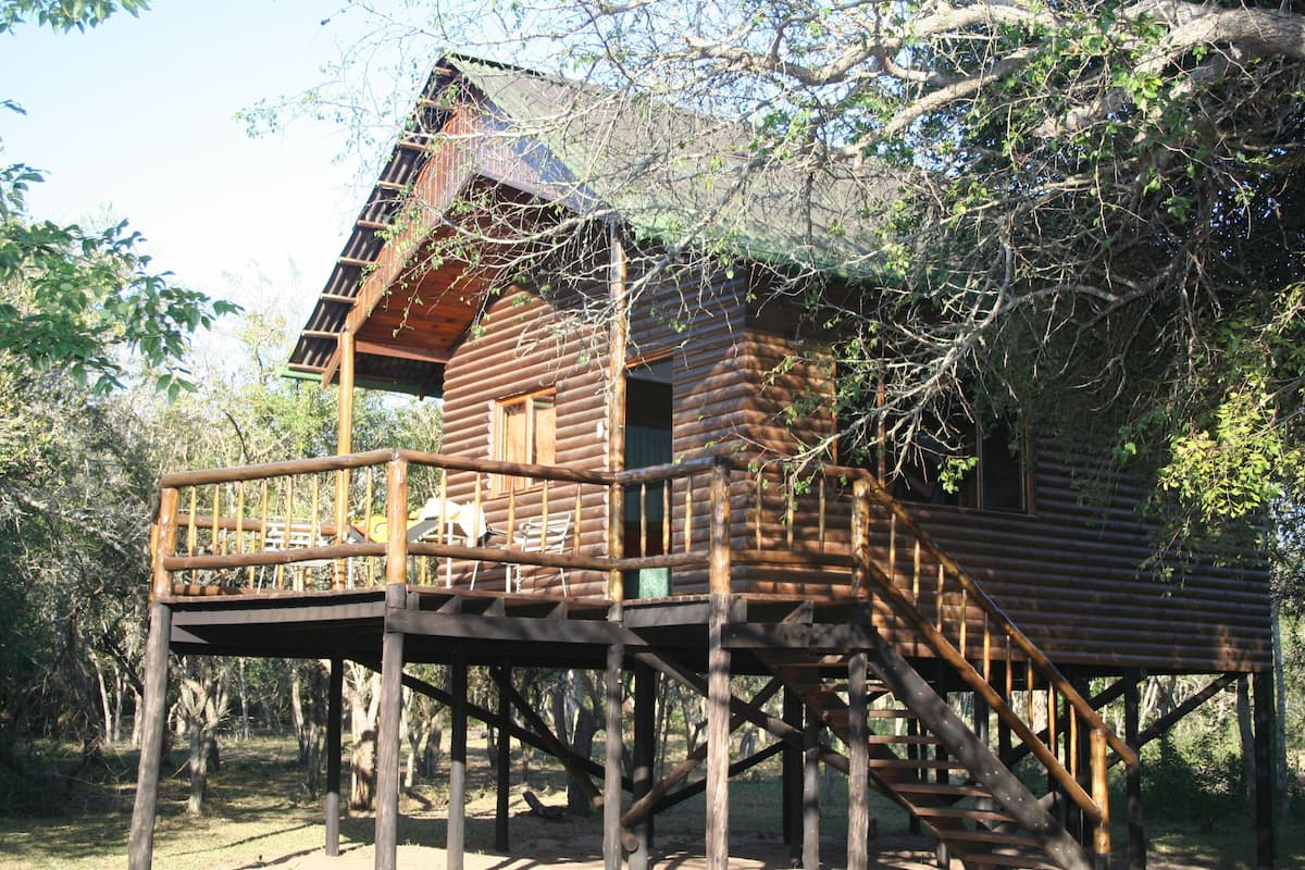 A wooden cabin with a balcony and stairs, surrounded by trees.