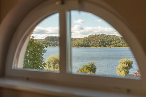 A view through a window with a semi-circular frame, overlooking a lake surrounded by trees and a forested hill. - Hotell Bogesund (Ulricehamn)
