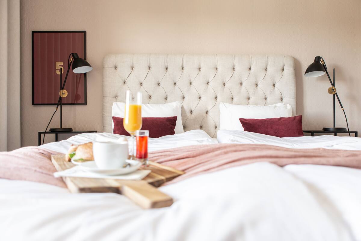 Cama bem arrumada com um edredom branco, duas almofadas vermelhas e uma bandeja de café da manhã com uma xícara, um copo de suco de laranja e um sanduíche.