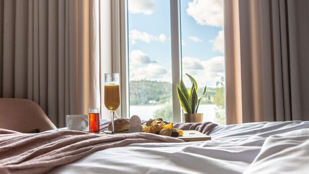 A bed with a view of a lake, a glass of orange juice, a cup, and a plate of fruit. - Hotell Bogesund (Ulricehamn)