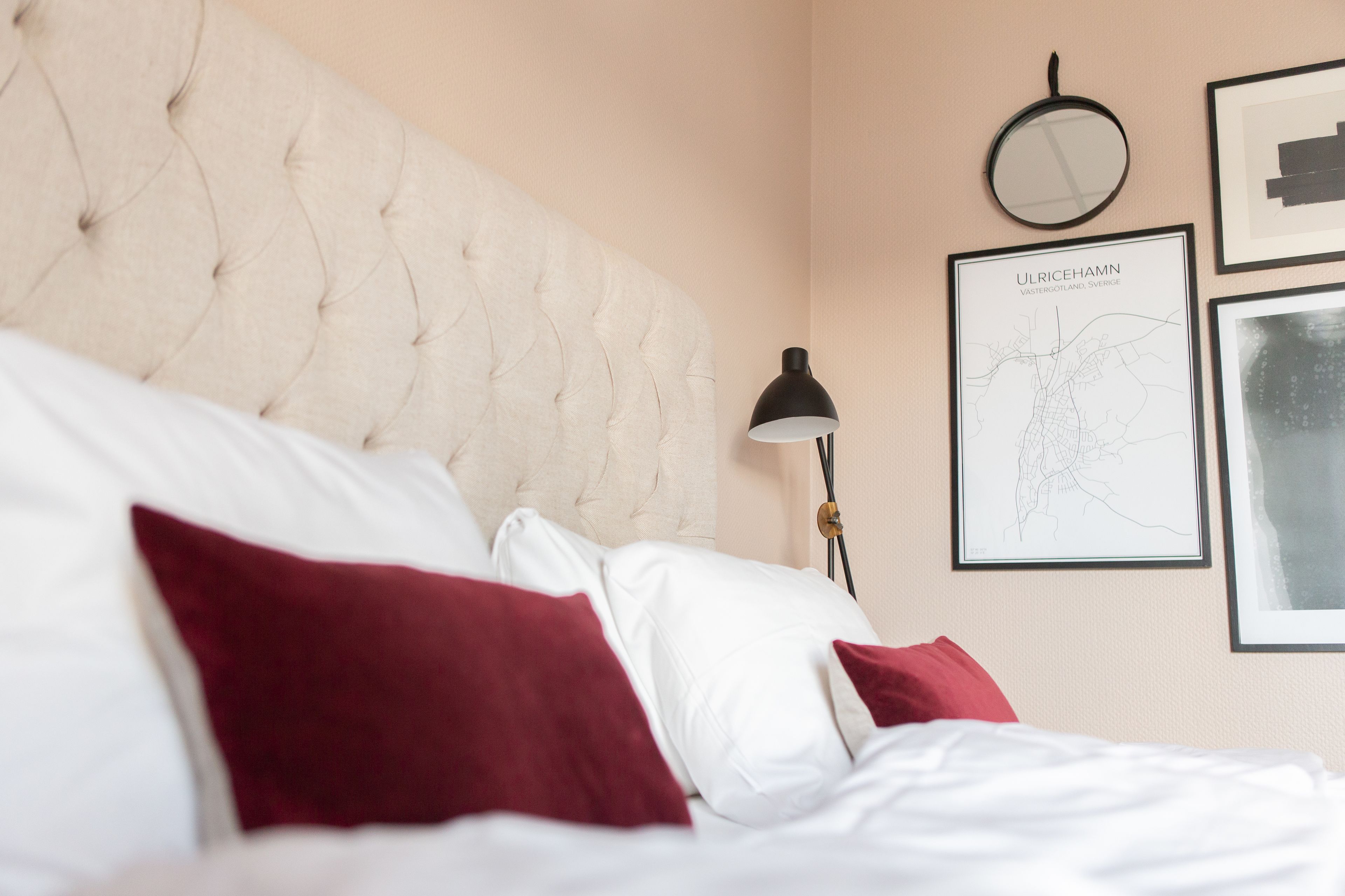 Una cama bien hecha con almohadas blancas y rojas, un cabecero capitonado, una lámpara de noche y una obra de arte enmarcada en la pared.