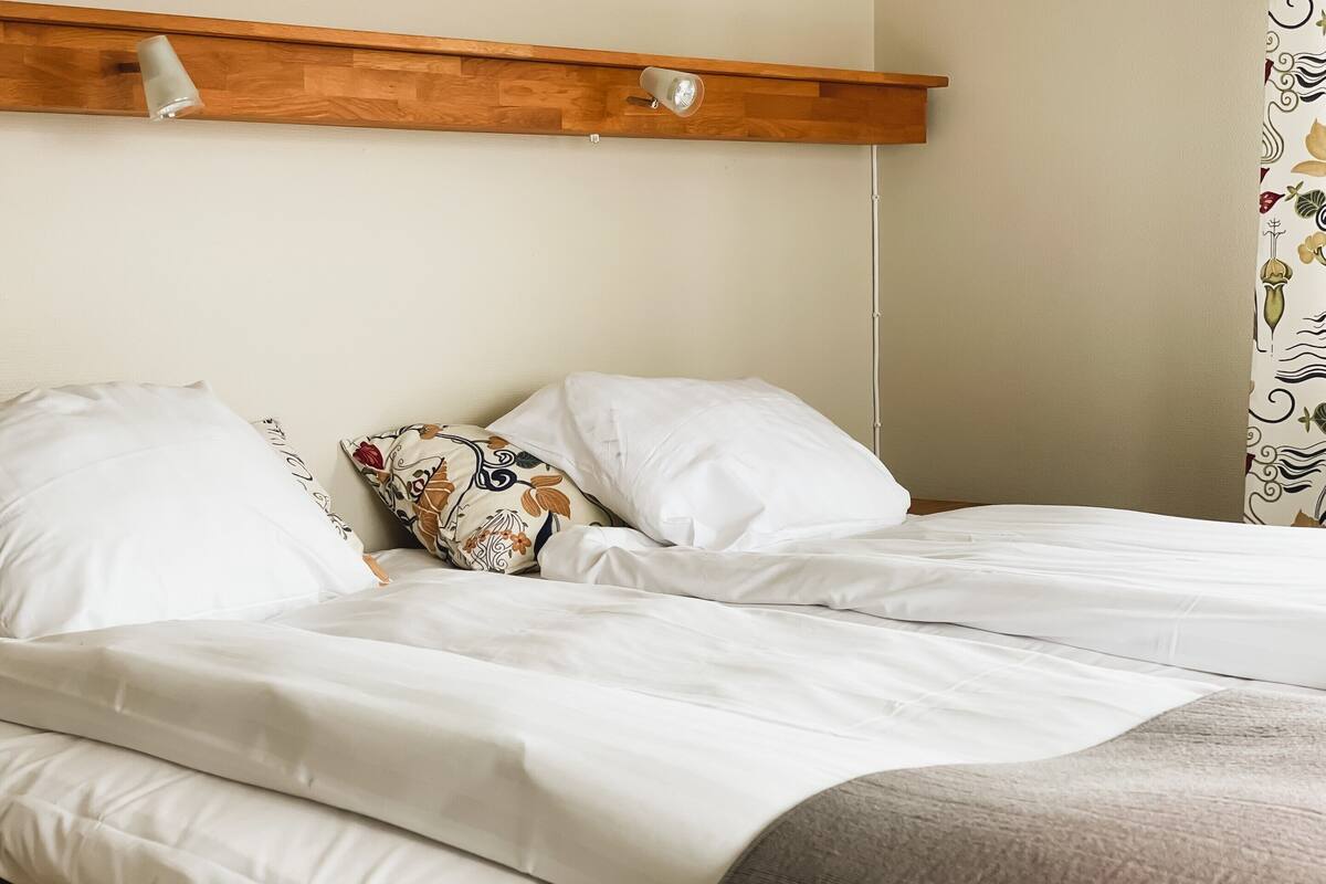 A double bed with white bedding and a decorative pillow, a wooden headboard with a light fixture, and a patterned curtain.
