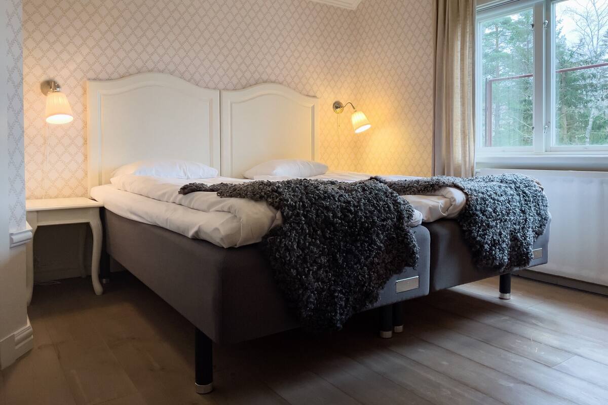 A neatly made bed with a dark blanket, two bedside lamps, and a window with curtains.