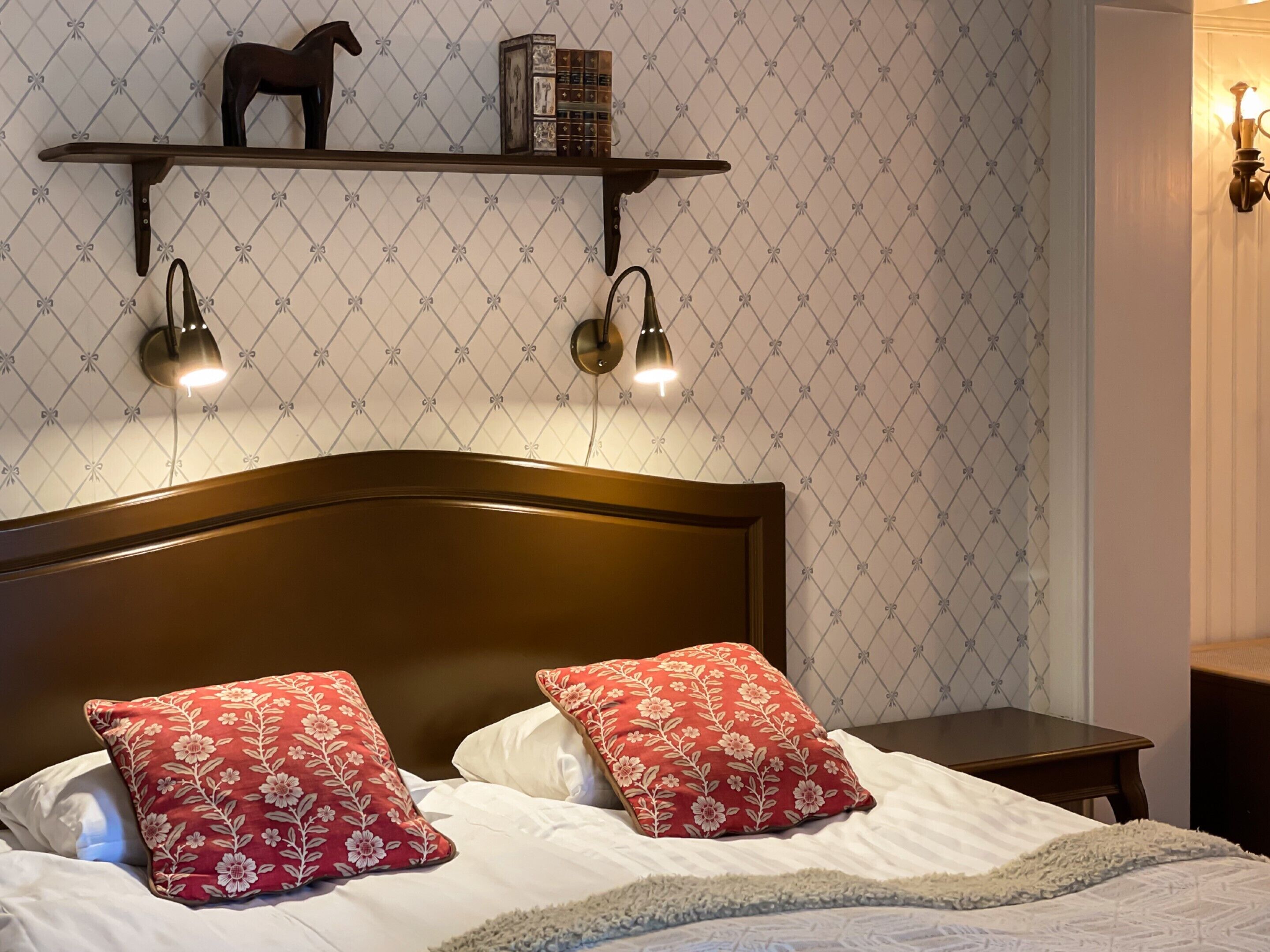 A neatly made bed with floral pillows, a wooden headboard, and a shelf above with decorative items and wall-mounted lights.