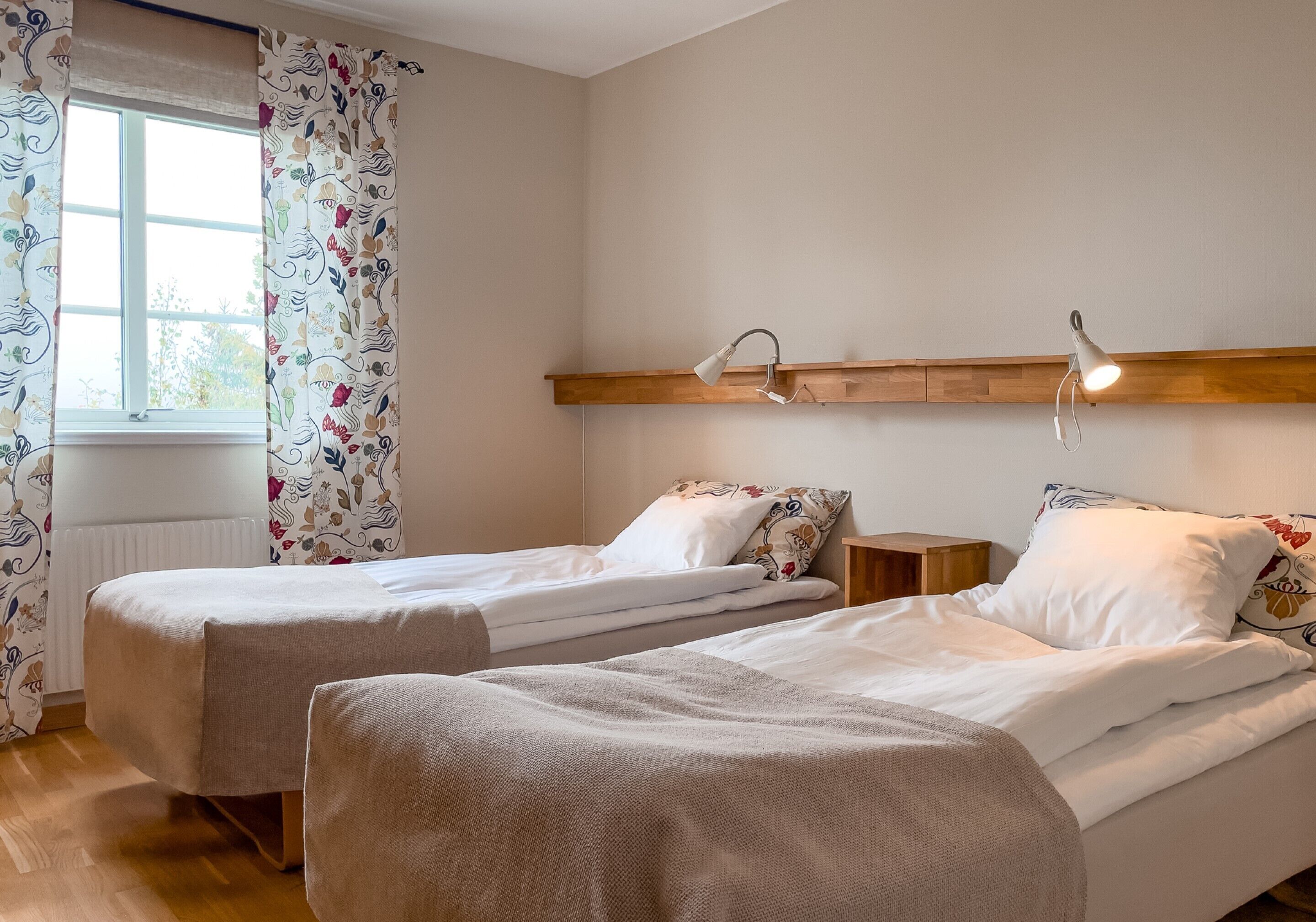 Quarto com duas camas de solteiro, uma janela com cortinas florais, uma prateleira de madeira com um abajur e um radiador.