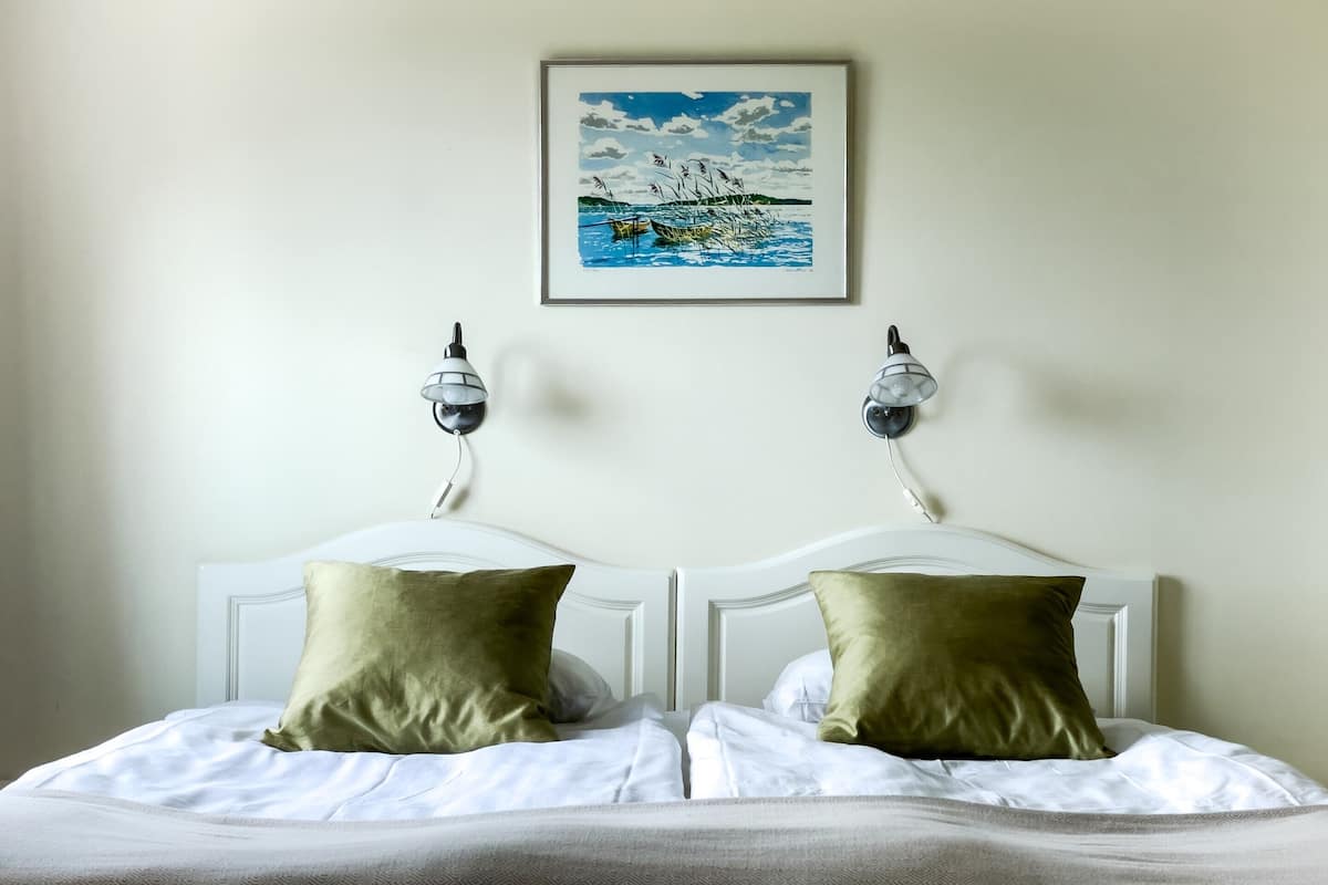 A double bed with two pillows, a framed painting of a boat on water, and two wall-mounted lamps.