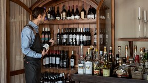 A bar area with a wooden wine rack filled with various bottles, a bartender in uniform, and a selection of liquor bottles on the counter. - Villa Långbers (Tallberg)