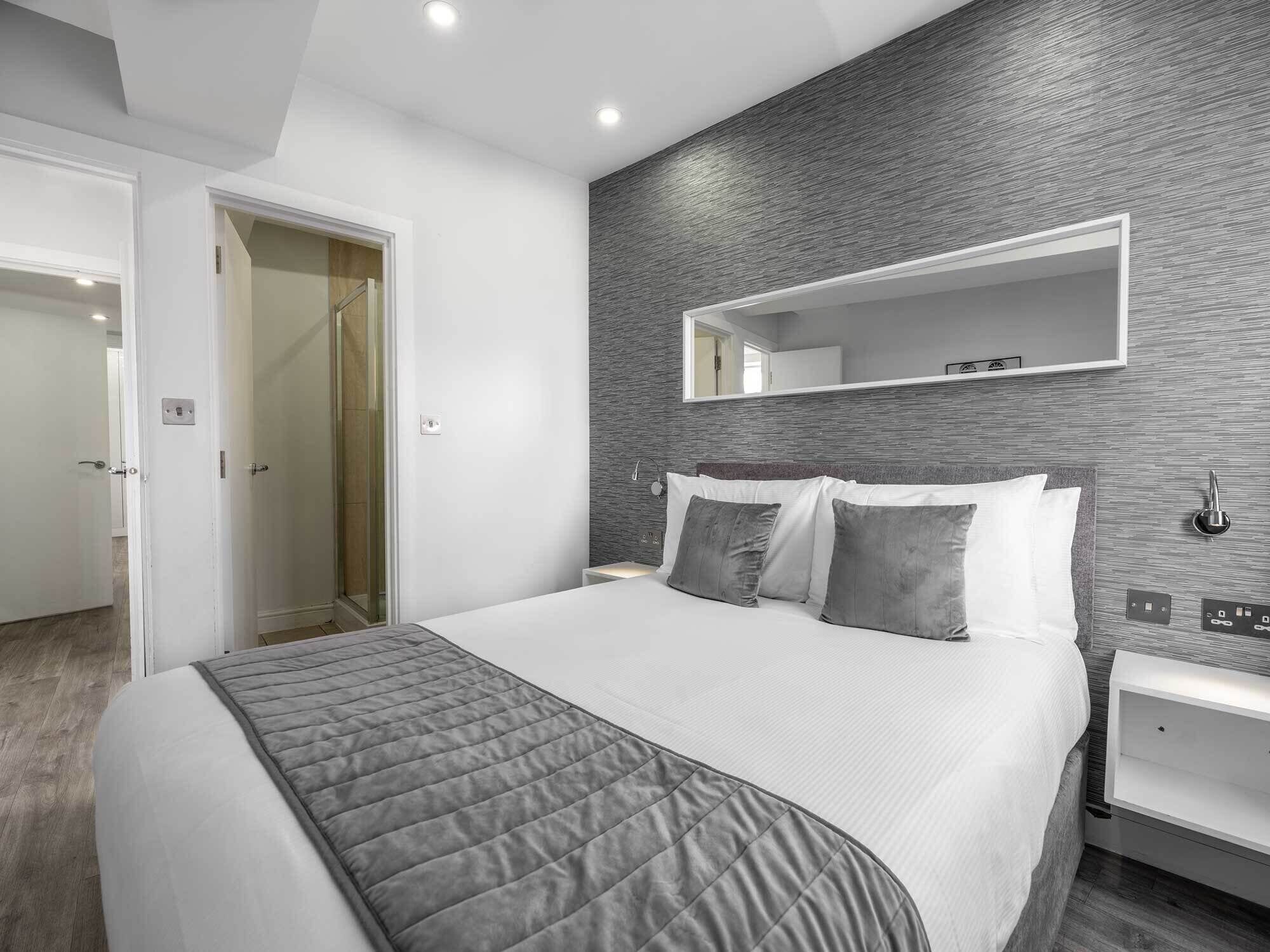 A modern bedroom with a large bed, a bedside table, a nightstand, a mirror, and a door leading to another room.
