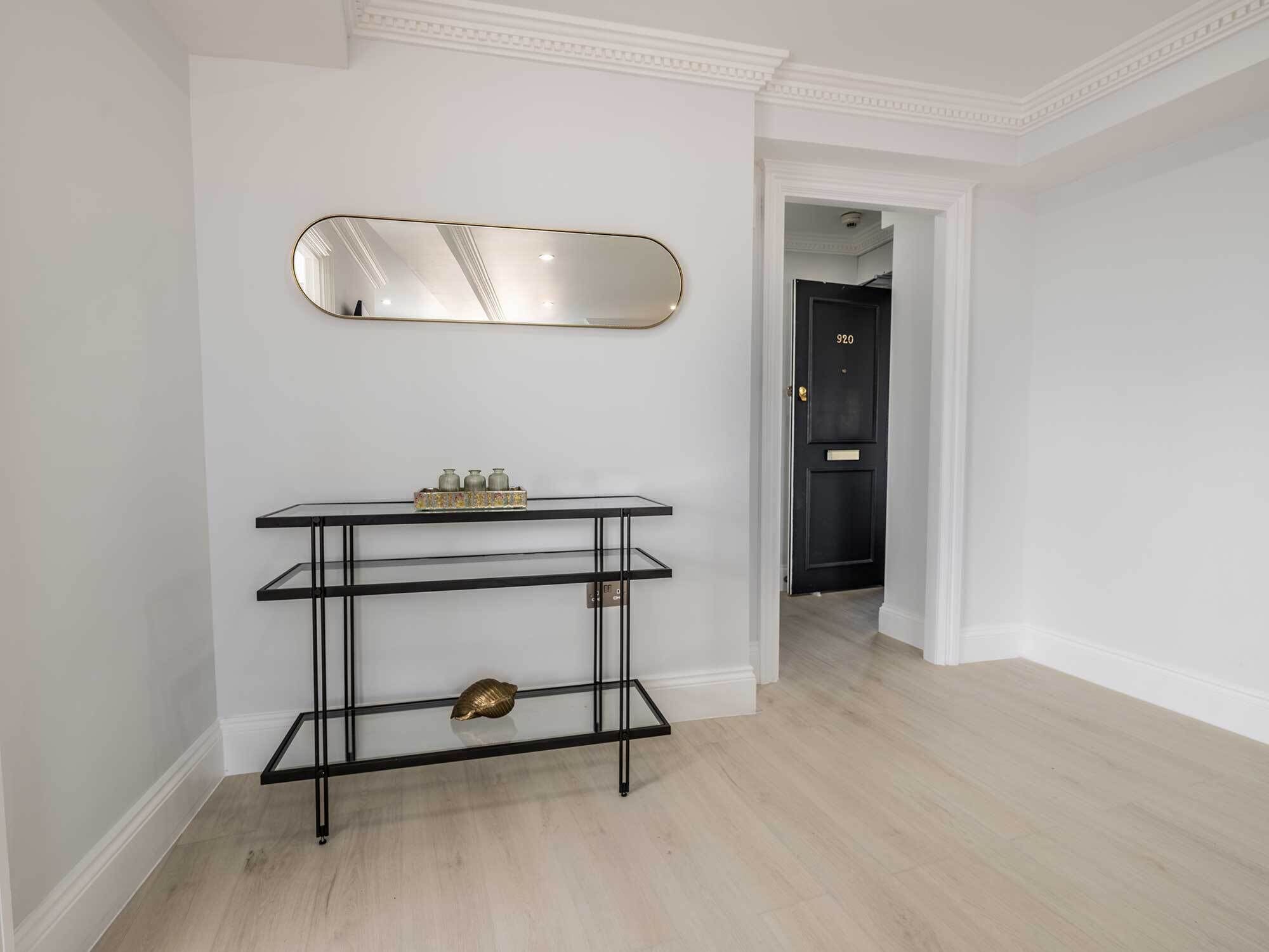A hallway with a black door marked "920", a white wall with a gold-framed mirror, and a black metal console table with decorative items.