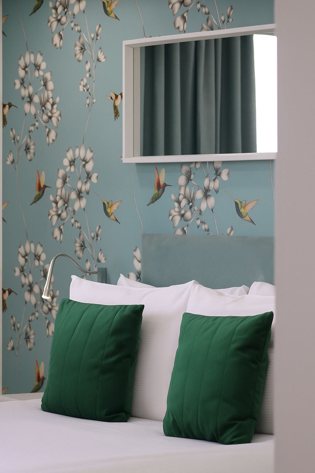 Een slaapkamer met een bed, groene kussens, een raam met gordijn en een wand met bloemen- en vogelmotief.
