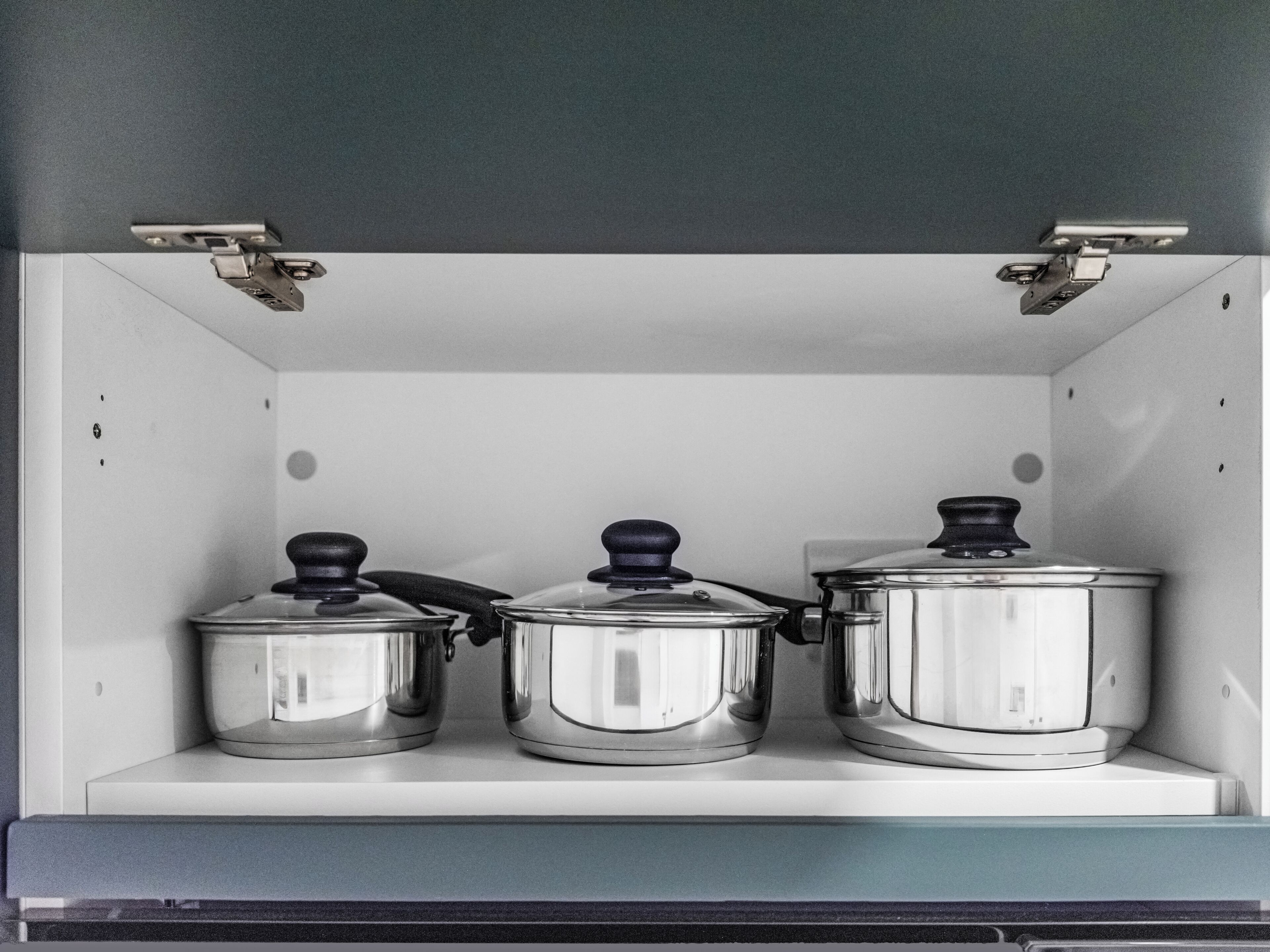 A kitchen cabinet with three stainless steel pots on a white shelf.