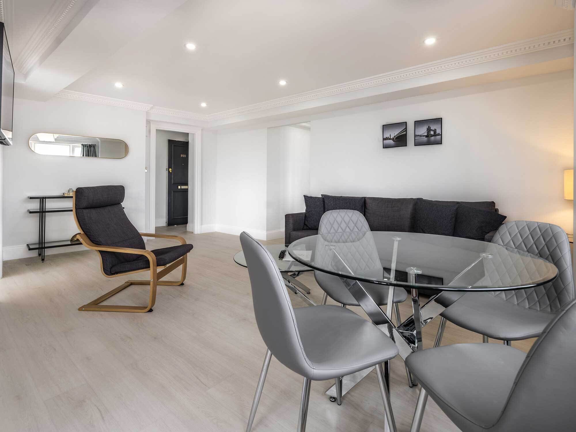 Een moderne woonkamer met een ronde glazen tafel, grijs gestoffeerde stoelen, een zwarte bank en een fauteuil met een zwart frame.