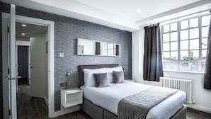 A modern bedroom with a large bed, a bedside table, a nightstand, a mirror, and a window with curtains. - Chelsea Cloisters Serviced Apartments (London)